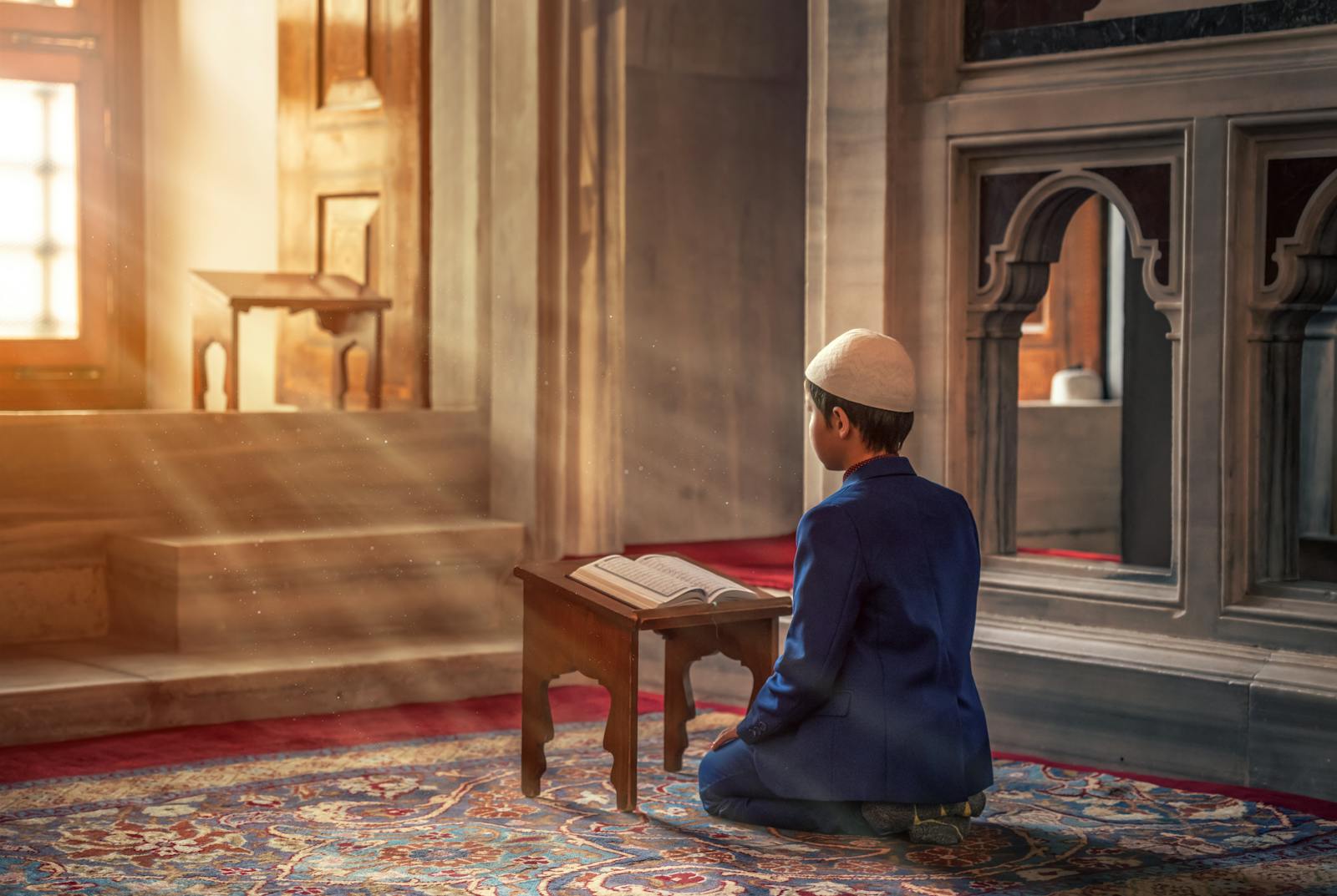 A young boy kneels in prayer inside a mosque with sunlight streaming through the window.